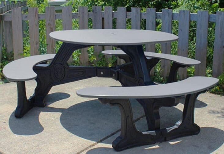 Bodega Outdoor Dining Table placed on a cement patio with a fence in the background. This table is made with our Green-Scapes designer frame which looks like black cast iron, but made from solid, recycled HDPE plastic. The top is made with HDPE sheet plastic. Shown with a black frame and gray top and seat.